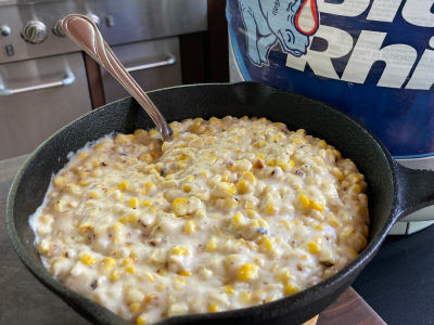 Completed cream corn mixture inside a cast iron skillet, sitting on a wooden table. Fresh Blue Rhino propane tank and standup propane grill in the background.